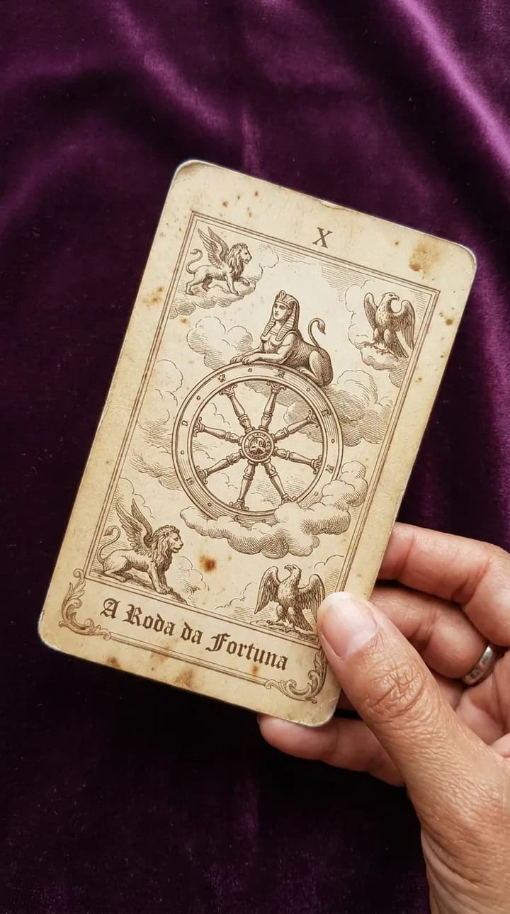 Mão segurando a carta A Roda da Fortuna, do baralho antigo, sobre o veludo roxo