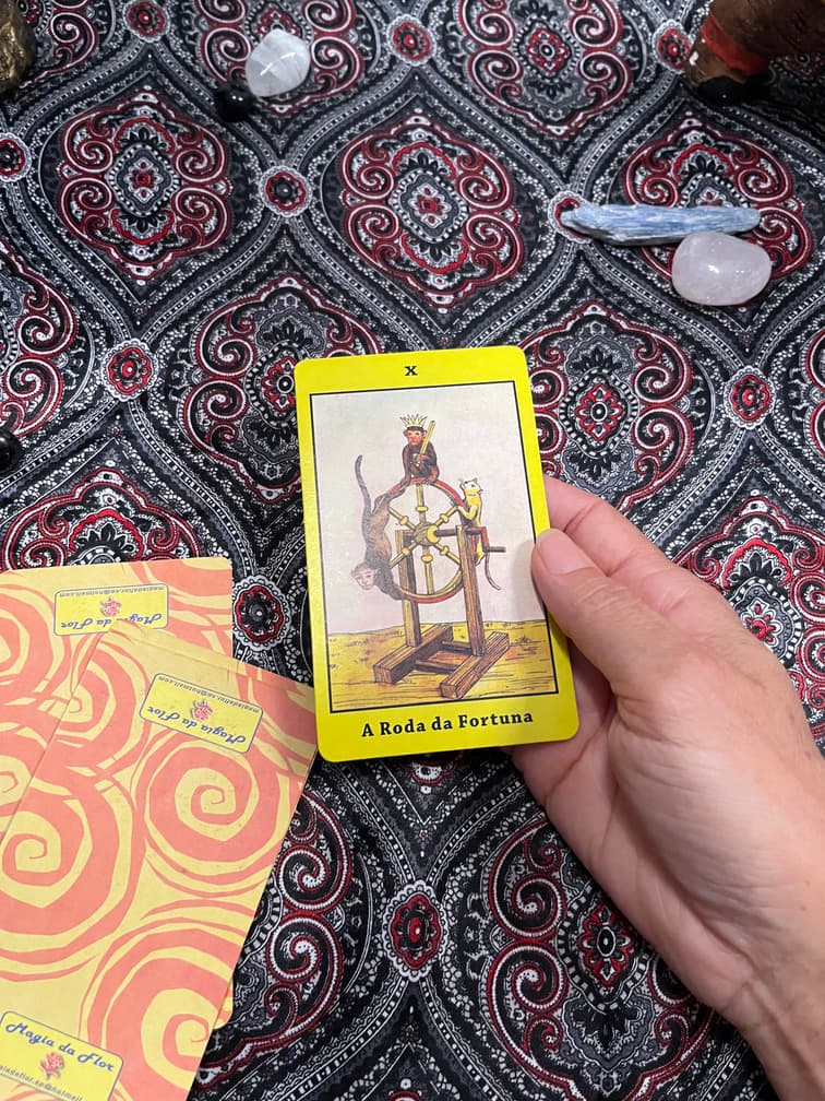 Mão segurando a carta A Roda da Fortuna sobre a toalha da mesa