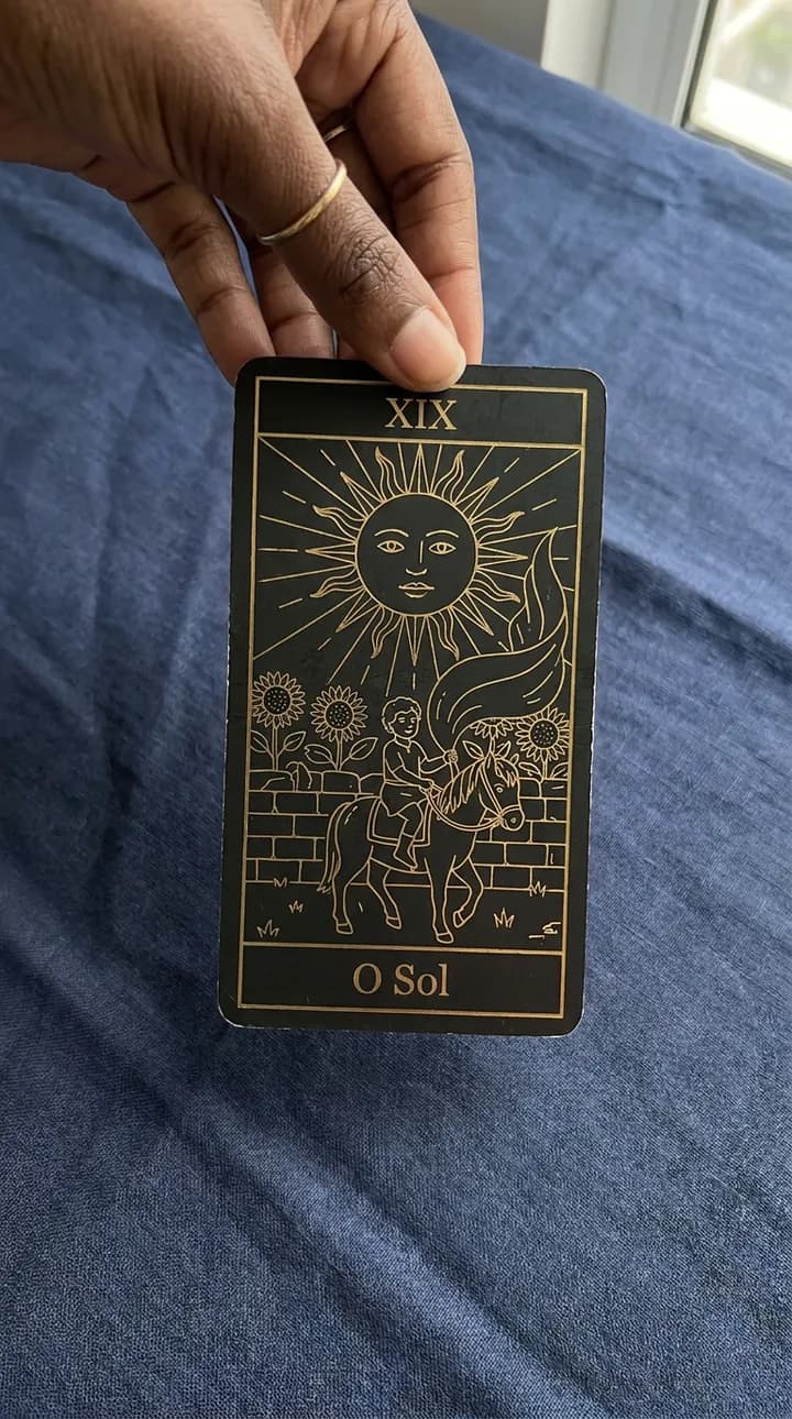 Hand holding The Sun card, from the black and gold deck, over the blue cloth